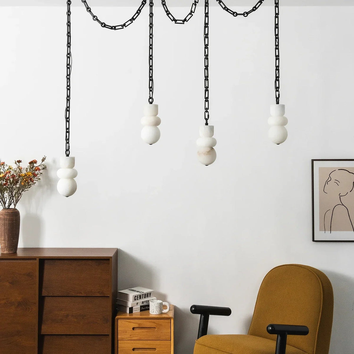 A white alabaster chandelier on black chains hangs above wooden drawers and a mustard armchair beside a framed line portrait.