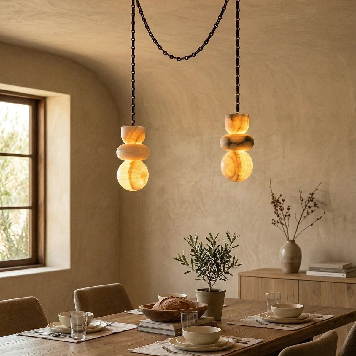 An alabaster chandelier with two glowing stone drops and draped black chains hangs above a set wooden dining table.