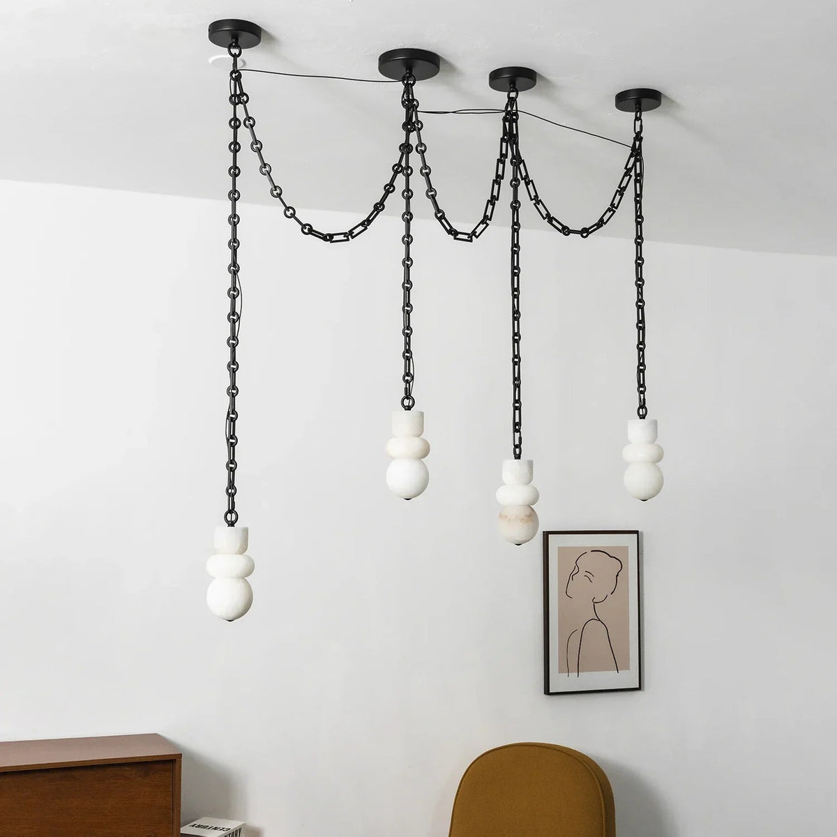 An unlit alabaster chandelier with four white sculpted shades and black chains hangs near a framed portrait and mustard chair.