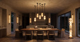 A modern dining room interior with a long wooden dining table, six upholstered chairs, a multi-light pendant chandelier, wall sconces, and a tall stacked floor lamp, all shown in a warm, atmospheric room setting.