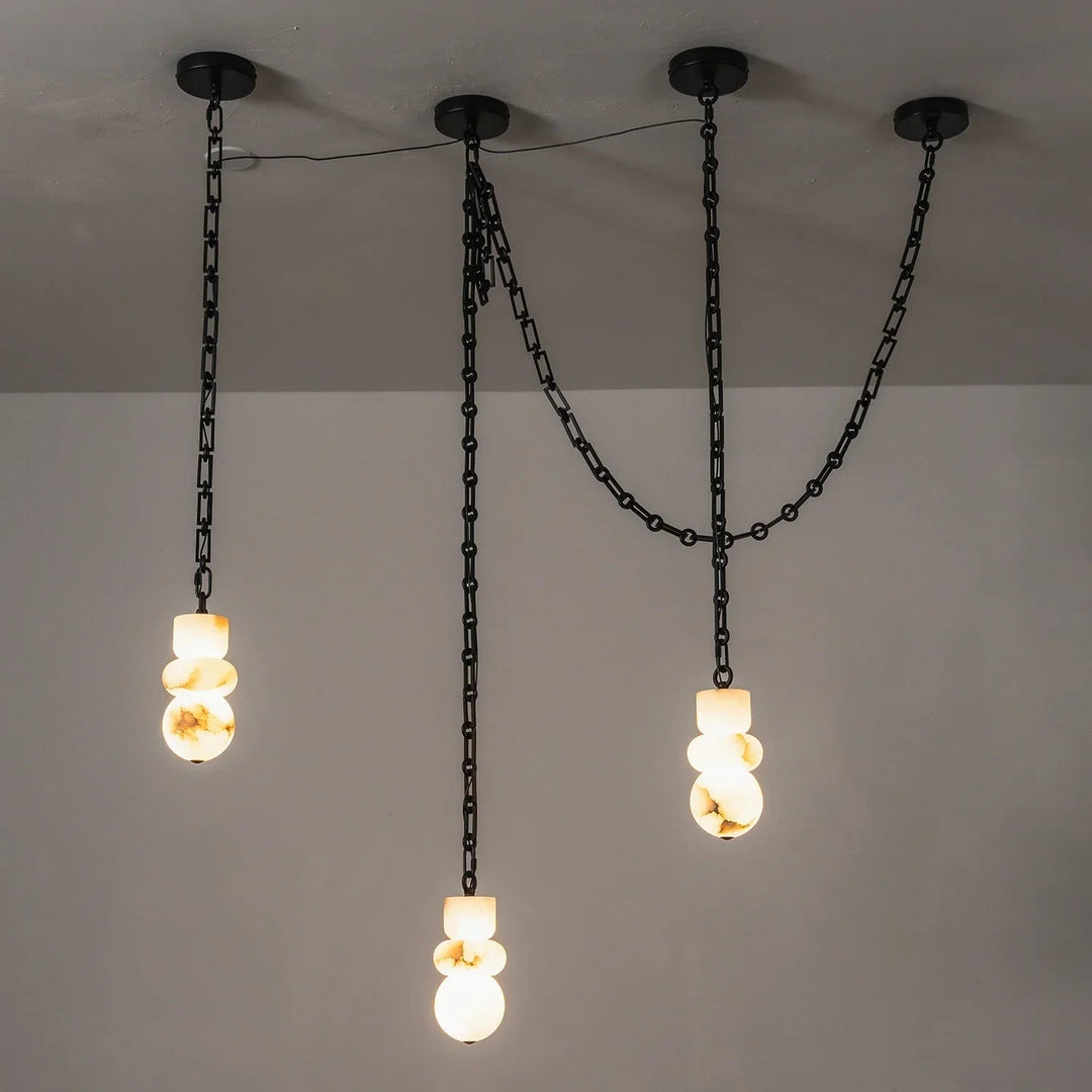 Alabaster chandelier with three glowing, veined stone shades suspended at staggered heights beneath black chains against a gray wall.