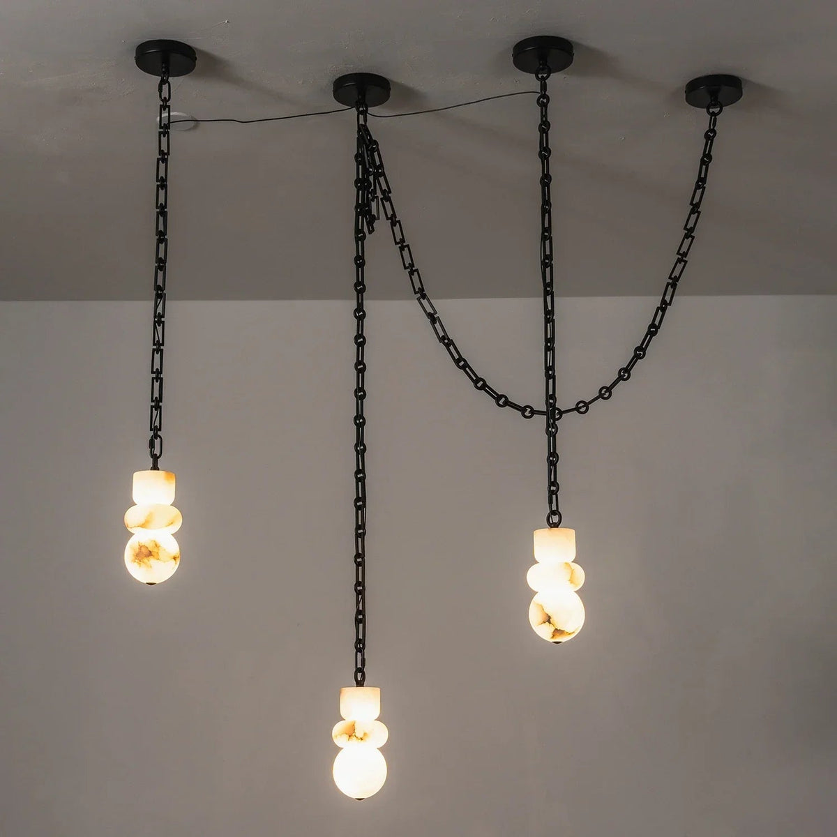 Alabaster chandelier with three glowing, veined stone shades suspended at staggered heights beneath black chains against a gray wall.