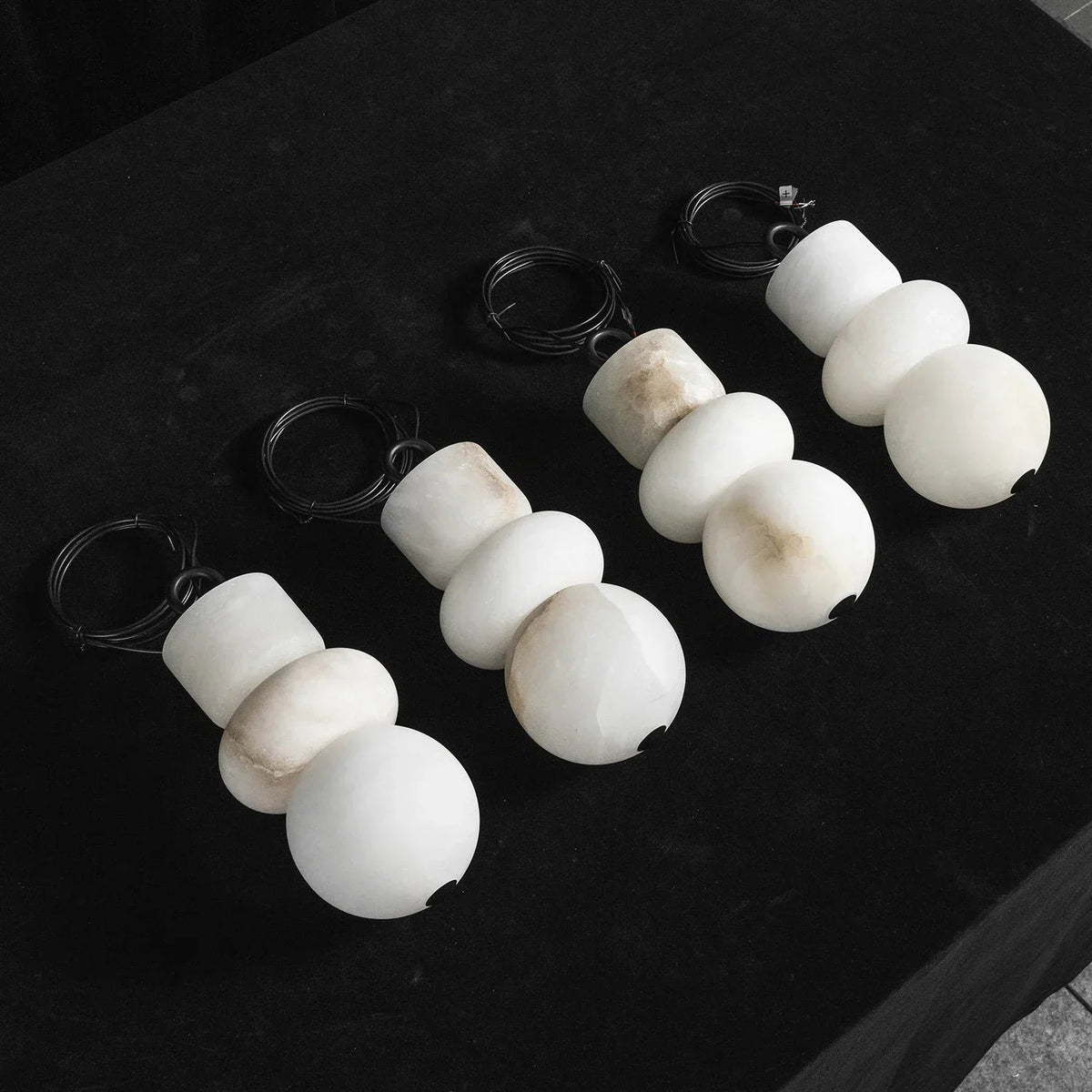 White alabaster chandelier shades in stacked sphere and cylinder shapes with coiled black cords on a dark surface.
