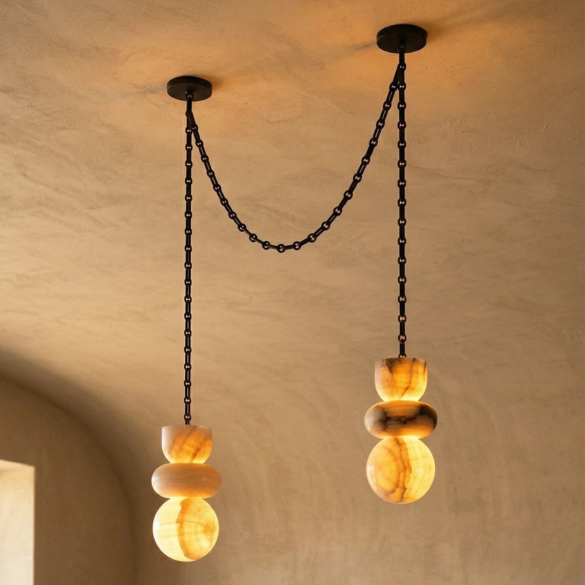 Alabaster chandelier with two glowing amber stone drops suspended at different heights from black chains against beige walls.