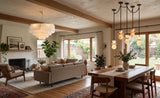 A modern, warm-toned living room and dining area featuring a large tiered white chandelier, a multi-light cluster pendant, a beige sofa, a wooden dining table, and large windows looking out to a backyard.