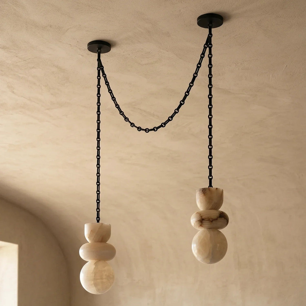 Unlit alabaster chandelier with two cream stone drops and draped black chains beneath a textured beige ceiling.