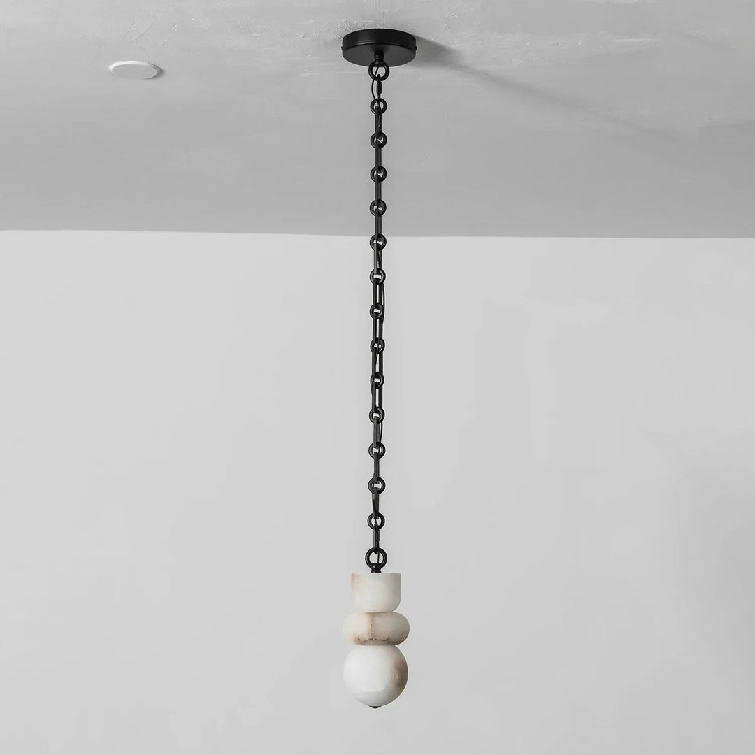 Unlit chandelier with stacked white alabaster shapes hangs from a straight black chain against a pale wall.