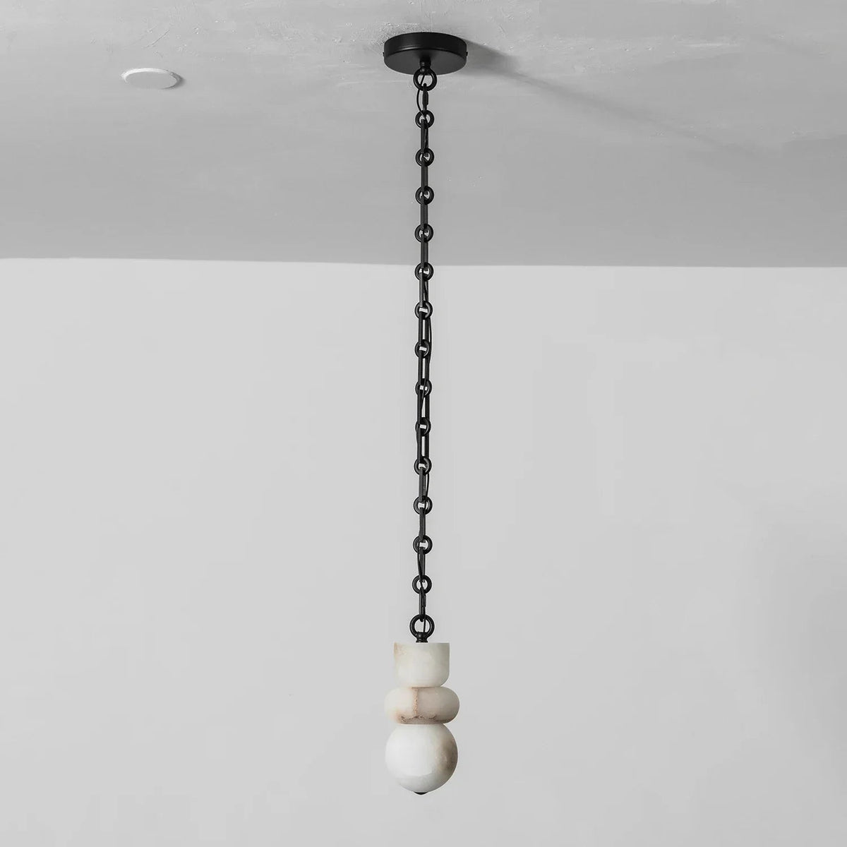 Unlit chandelier with stacked white alabaster shapes hangs from a straight black chain against a pale wall.