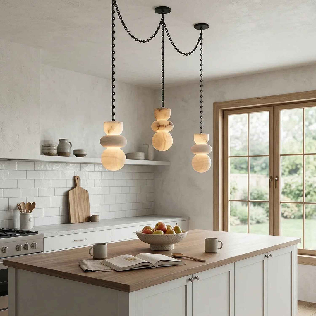 An alabaster chandelier with three stacked cream stone drops hangs above a wooden kitchen island beside large windows.