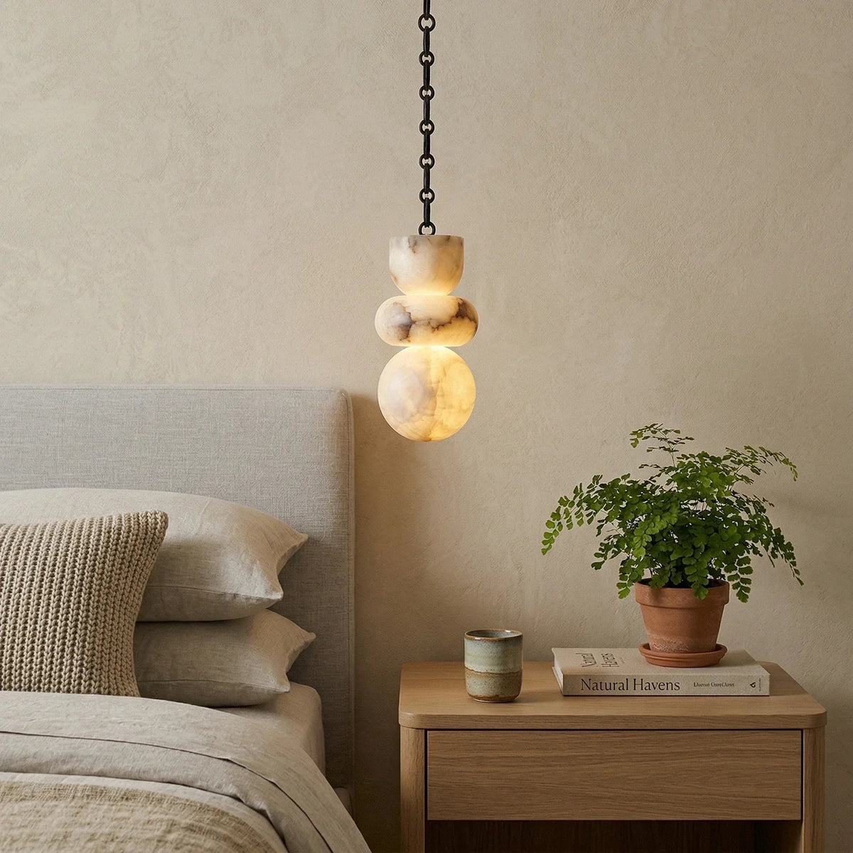 A cream alabaster chandelier with brown veining hangs on a black chain above a bedside table with a potted fern.