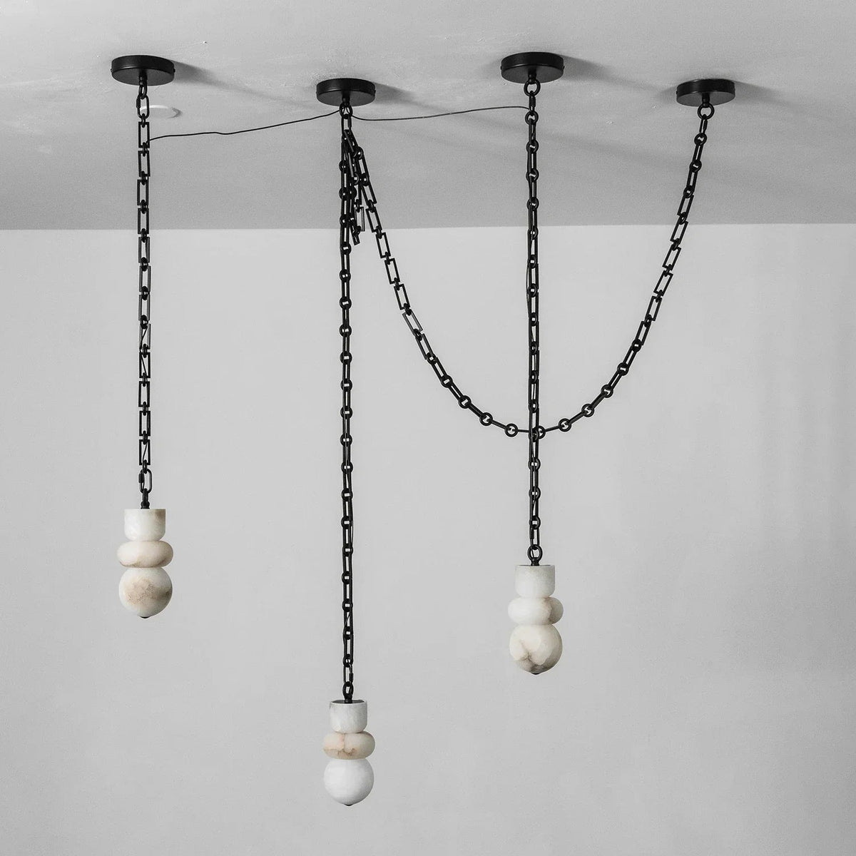 Unlit alabaster chandelier with three white sculpted shades, staggered black chains, and four round ceiling mounts against a pale background.
