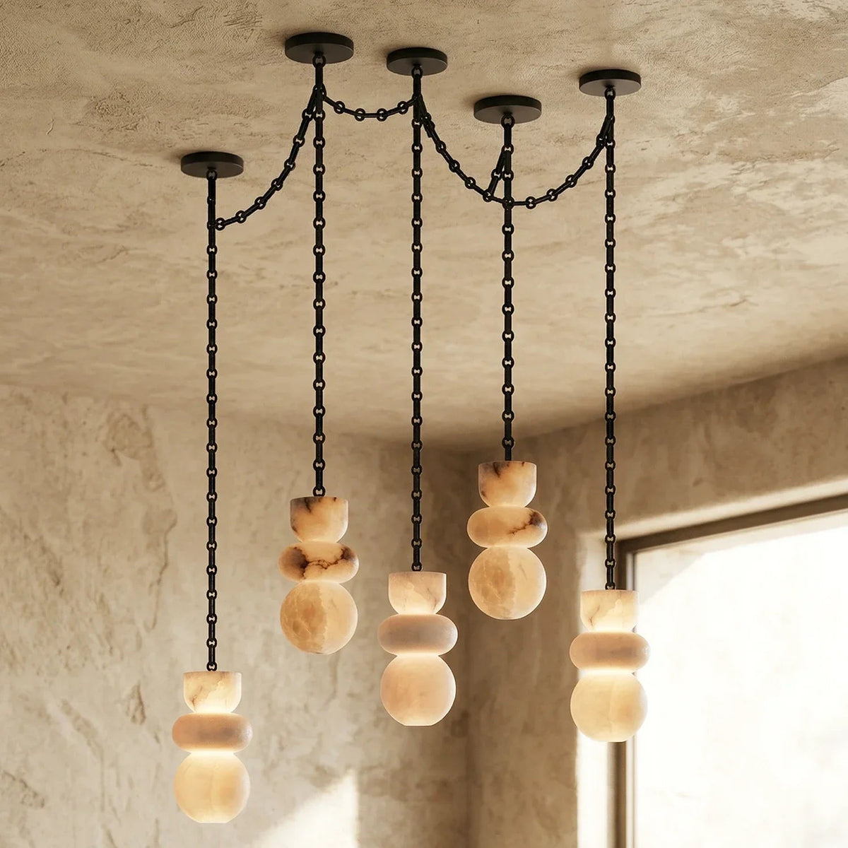 Alabaster chandelier with five illuminated cream stone drops suspended from black chains beside a window in a textured beige room.