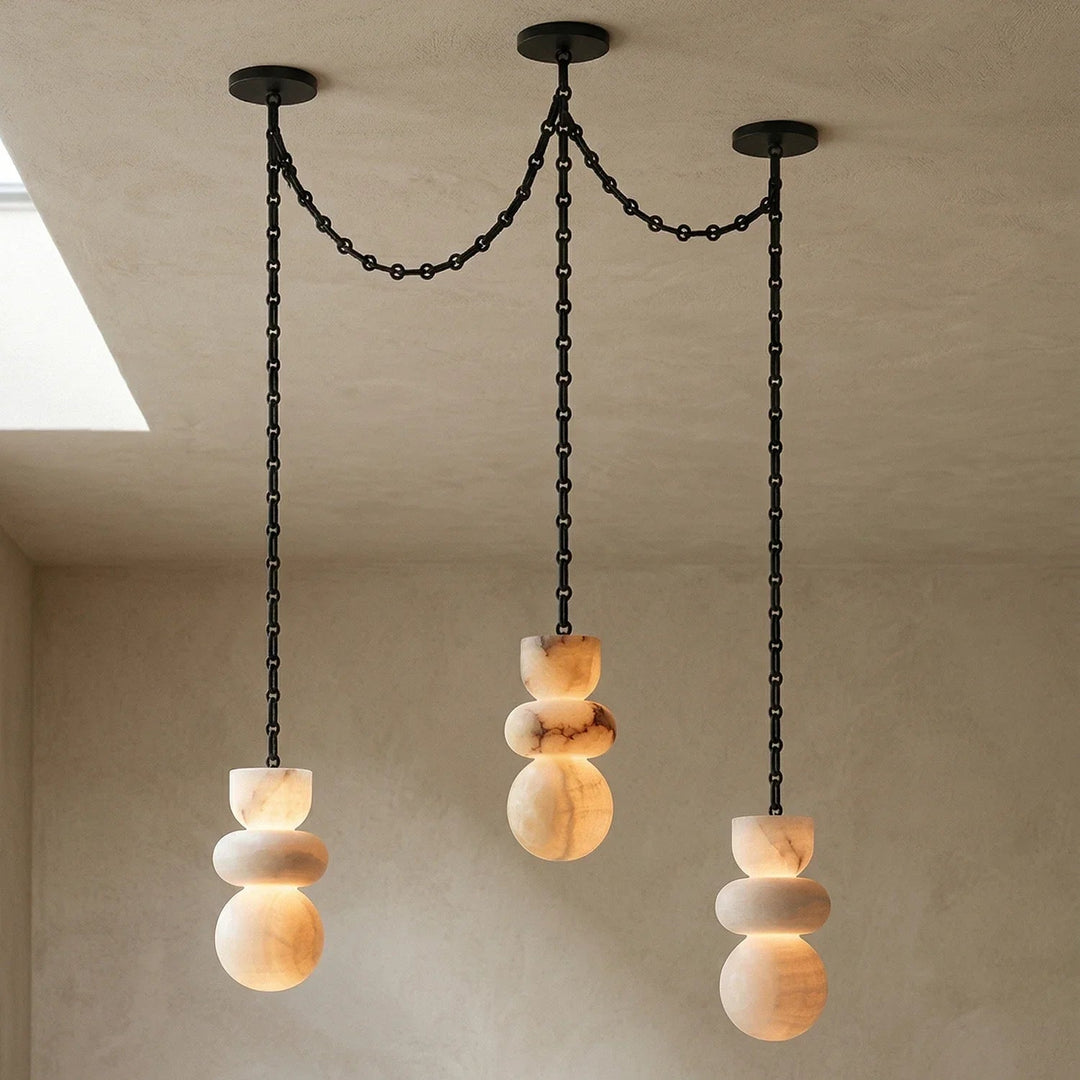 Alabaster chandelier with three illuminated cream stone drops hanging from connected black chains beside a ceiling opening.