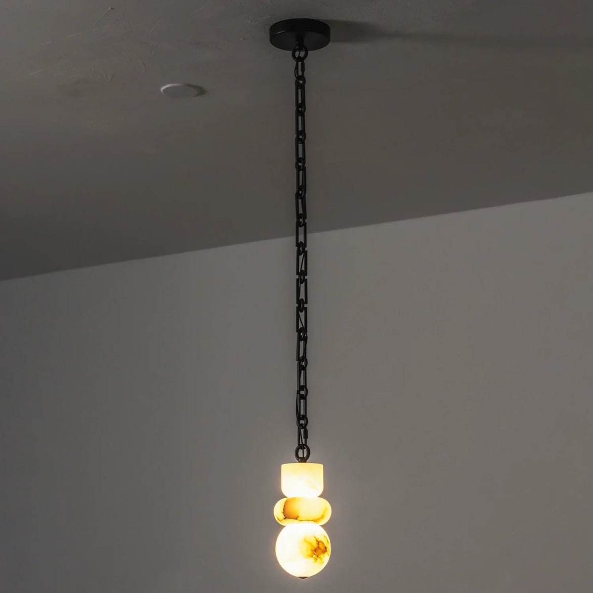 Alabaster chandelier glows warm yellow beneath a long black chain against a gray wall.