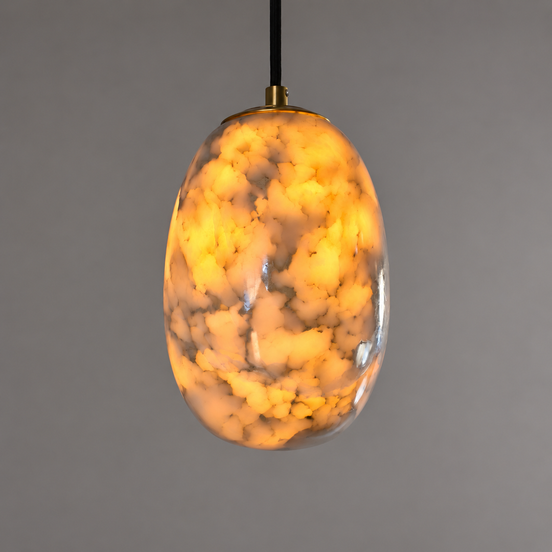 An oval pendant light with a glowing, marbled amber and cream glass shade, suspended by a black cord with brass hardware against a solid gray background.