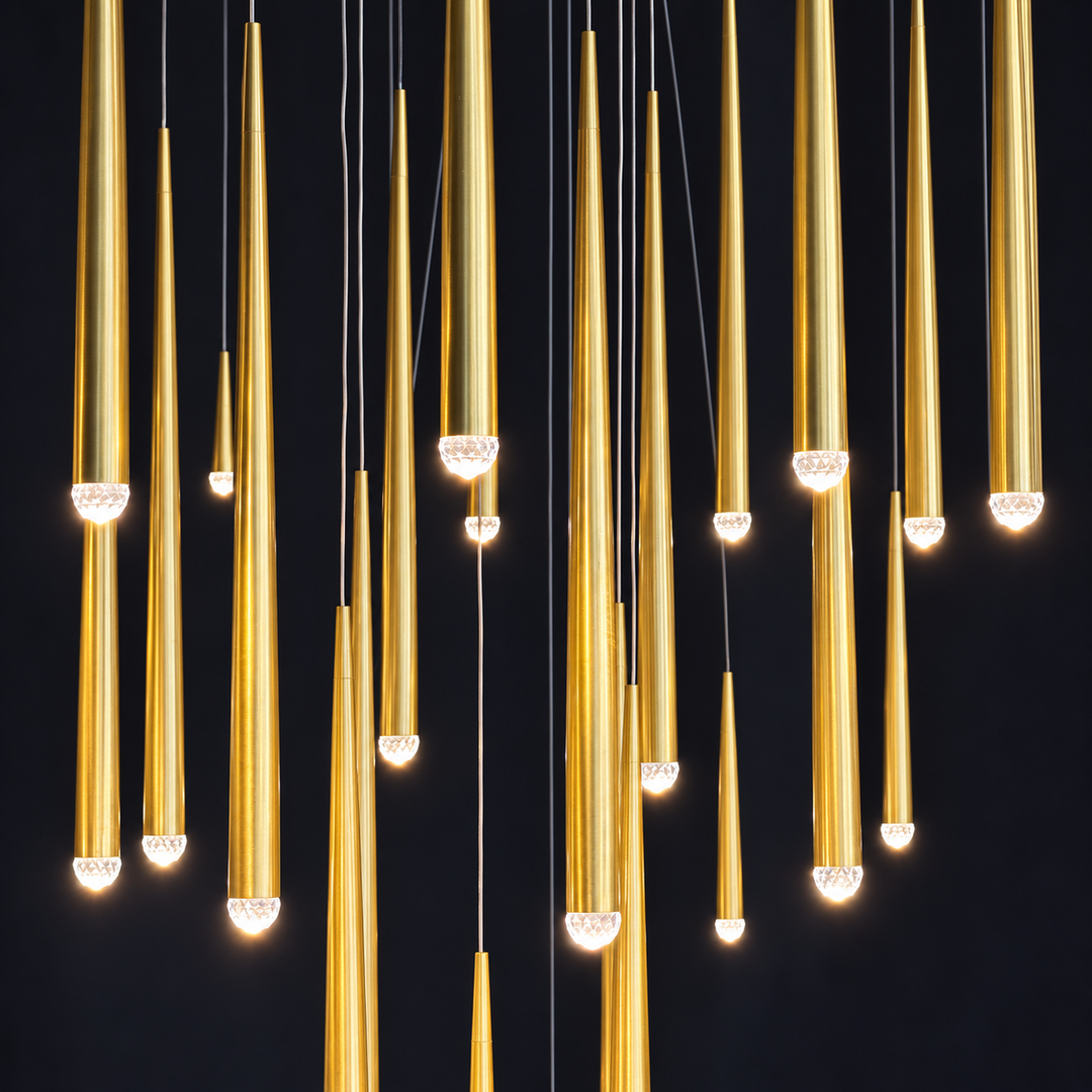 A modern chandelier featuring multiple tapered gold metal rods of varying lengths, suspended from thin black wires against a solid black background. Each rod is illuminated at the bottom with a clear faceted crystal diffuser that glows with a warm light.