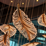 A close-up of a large hanging leaf-shaped chandelier made of textured clear glass with warm metallic gold-toned edges, suspended by thin cables in an indoor setting with blurred architectural background.