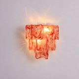 A sculptural wall sconce featuring layered, translucent red-orange glass panels that glow warmly from an internal light source, mounted against a plain light gray wall.