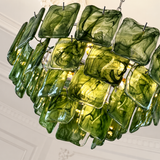 A modern chandelier featuring multiple square glass panels in a marbled olive green and yellow-green finish, suspended in a curved arrangement against a white paneled ceiling.
