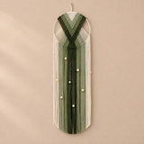 A vertical macrame wall hanging with a geometric chevron pattern in shades of green and cream, adorned with small white beads, hanging on a beige wall.