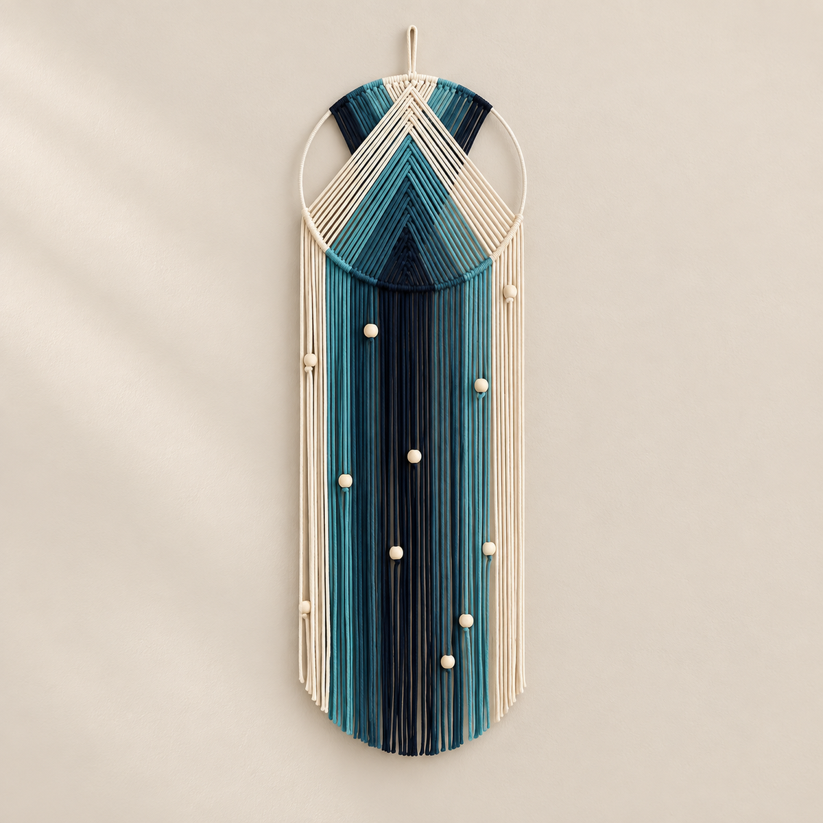 A bohemian-style macrame wall hanging featuring a geometric pattern in shades of teal, blue, and black, suspended from off-white cords with small decorative beads, shown hanging on a neutral beige wall.