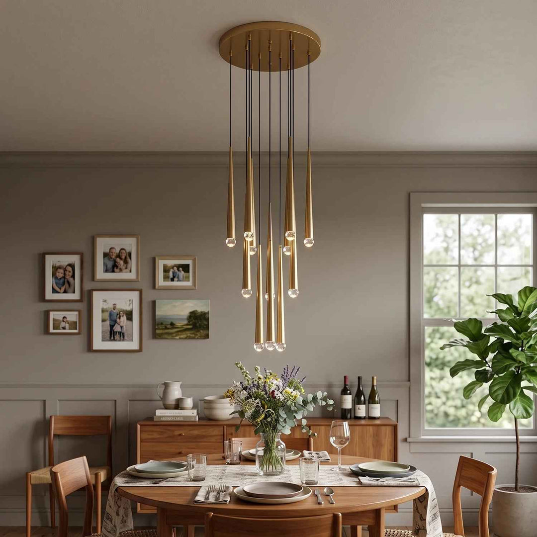 A modern multi-light linear chandelier with warm brass-toned tubes and clear glass bulbs hangs over a wooden dining table in a styled dining room with gray walls, framed photos, and a window.