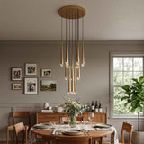 A modern multi-light linear chandelier with warm brass-toned tubes and clear glass bulbs hangs over a wooden dining table in a styled dining room with gray walls, framed photos, and a window.
