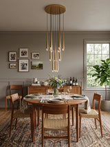 A modern dining room featuring a round wooden dining table surrounded by six woven-back chairs, under a multi-light chandelier. The room has gray walls, a patterned rug, and a wooden sideboard in the background.