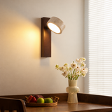 A modern wall-mounted sconce with a cylindrical textured concrete shade and a dark wood backplate, casting a warm yellow light on a beige wall above a wooden table with a vase of white flowers and fruit.