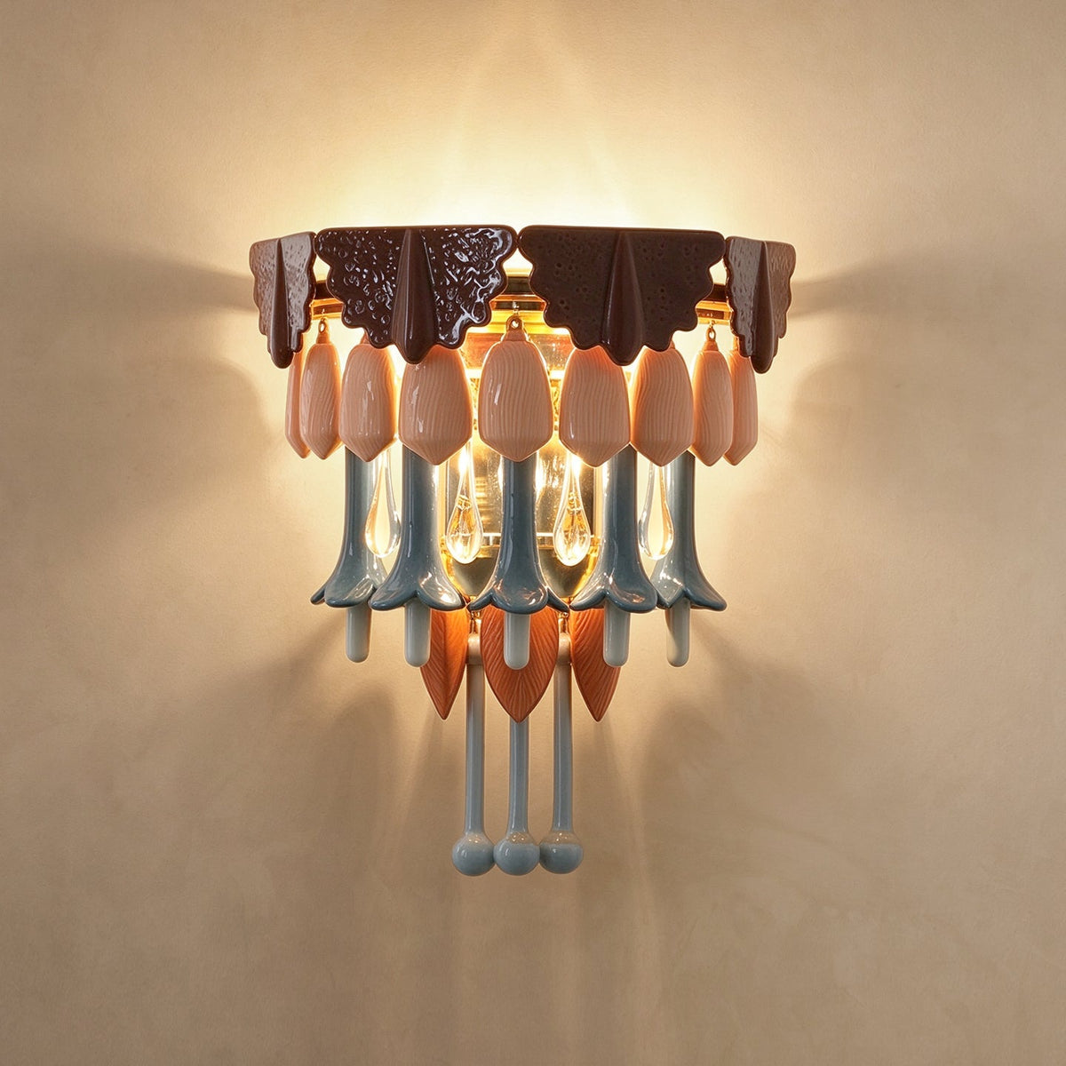 A decorative wall sconce light fixture with a dark brown top plate, pale pink teardrop-shaped glass shades, and light blue hanging glass elements, mounted on a beige wall and illuminated.