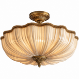 A semi-flush mount ceiling light fixture featuring a warm, cream-colored pleated glass shade and ornate antique brass metal hardware.