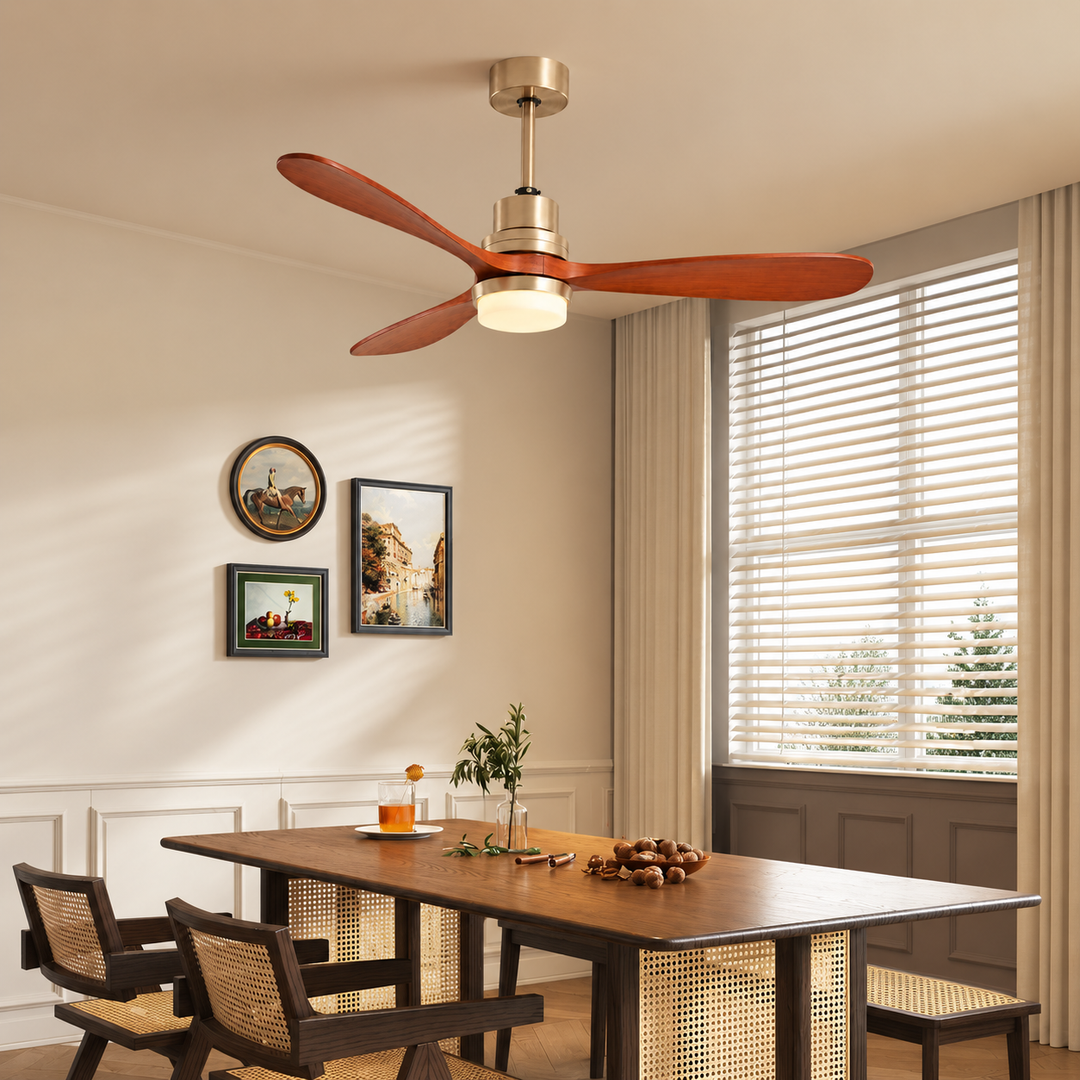 A ceiling fan with four reddish-brown wooden blades and a warm brass fixture housing a glowing light, installed in a dining room with a wooden table, woven chairs, and a window with blinds.