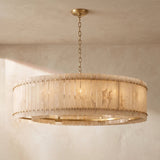 A large circular chandelier with a warm brass frame and a translucent beige shell shade, hanging from a chain in a textured beige room.