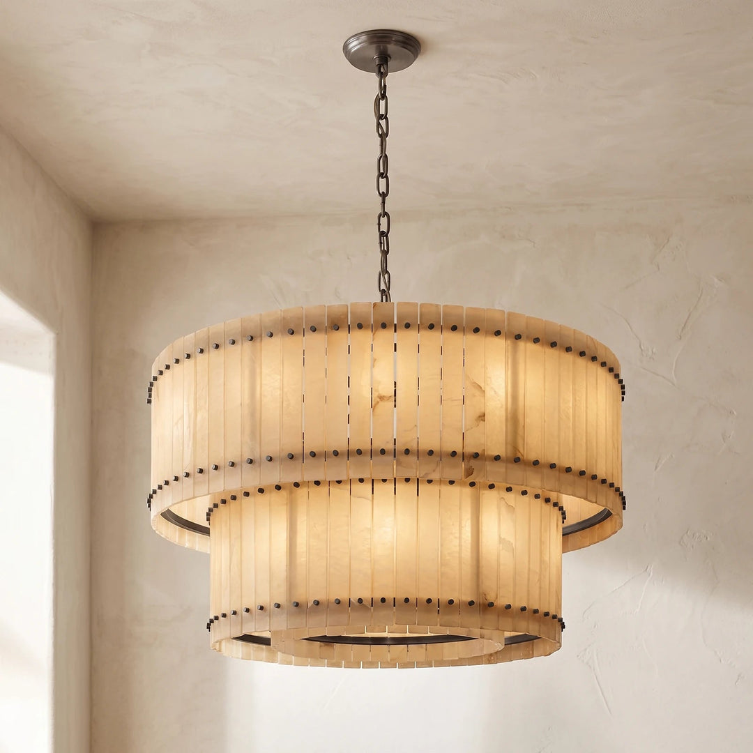 A two-tier drum pendant chandelier made from natural wood slats with dark metal rivets, hanging from a dark chain in a room with textured beige walls and ceiling.