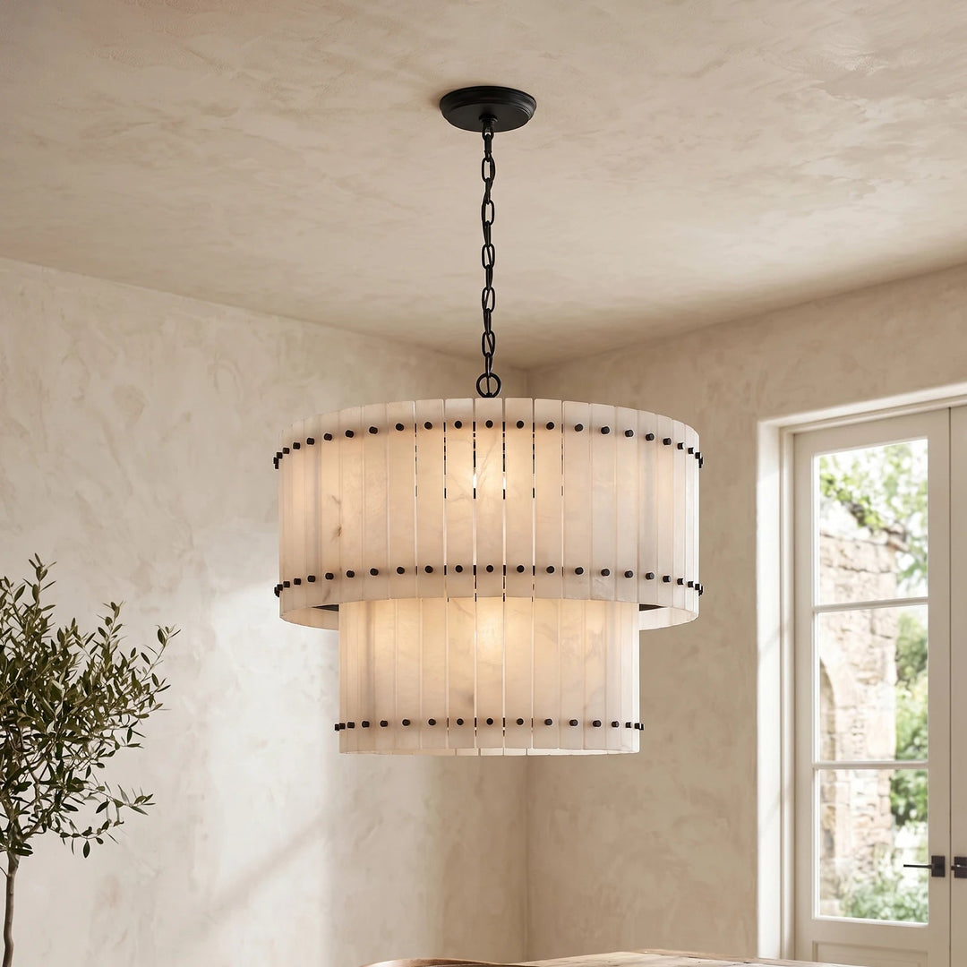 A two-tier drum pendant chandelier with a warm white pleated shade and black metal hardware, hanging in a room with textured beige walls and a window.