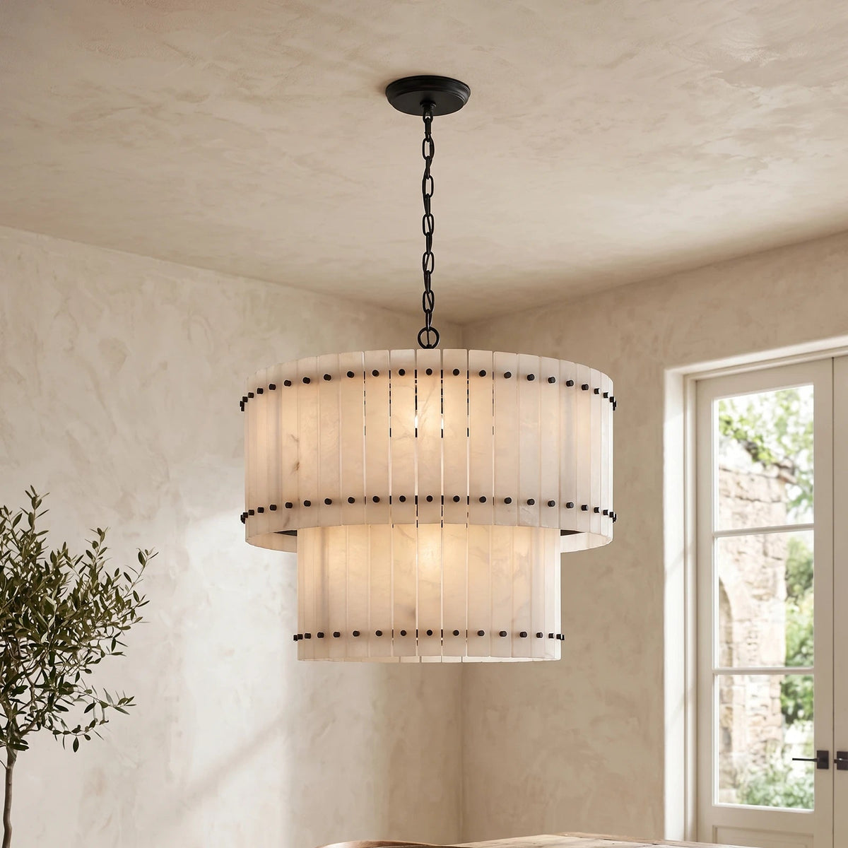 A two-tier drum pendant chandelier with a warm white pleated shade and black metal hardware, hanging in a room with textured beige walls and a window.