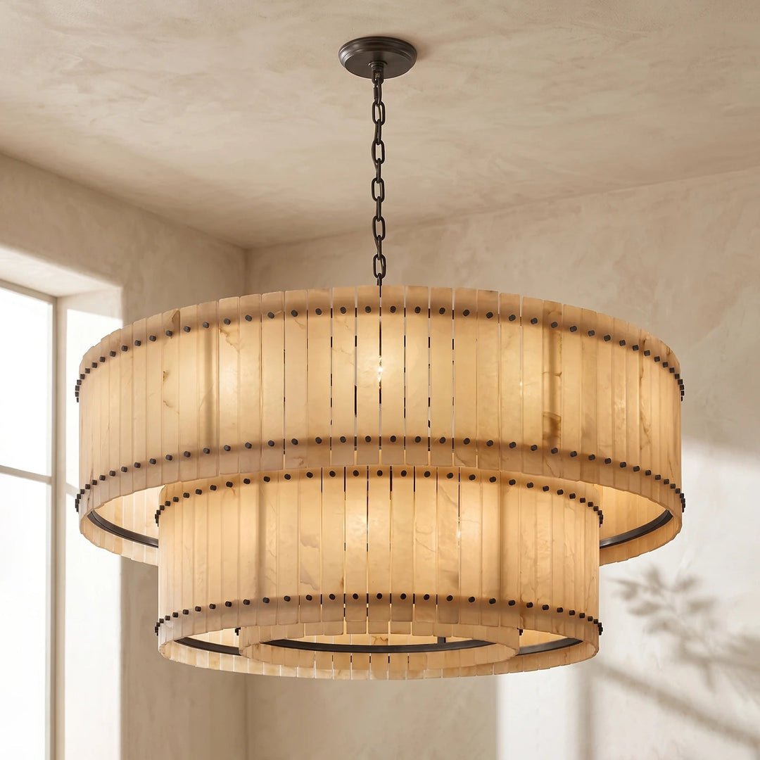 A two-tiered drum chandelier with a warm beige woven shade and dark metal hardware, hanging from a chain in a room with textured beige walls.