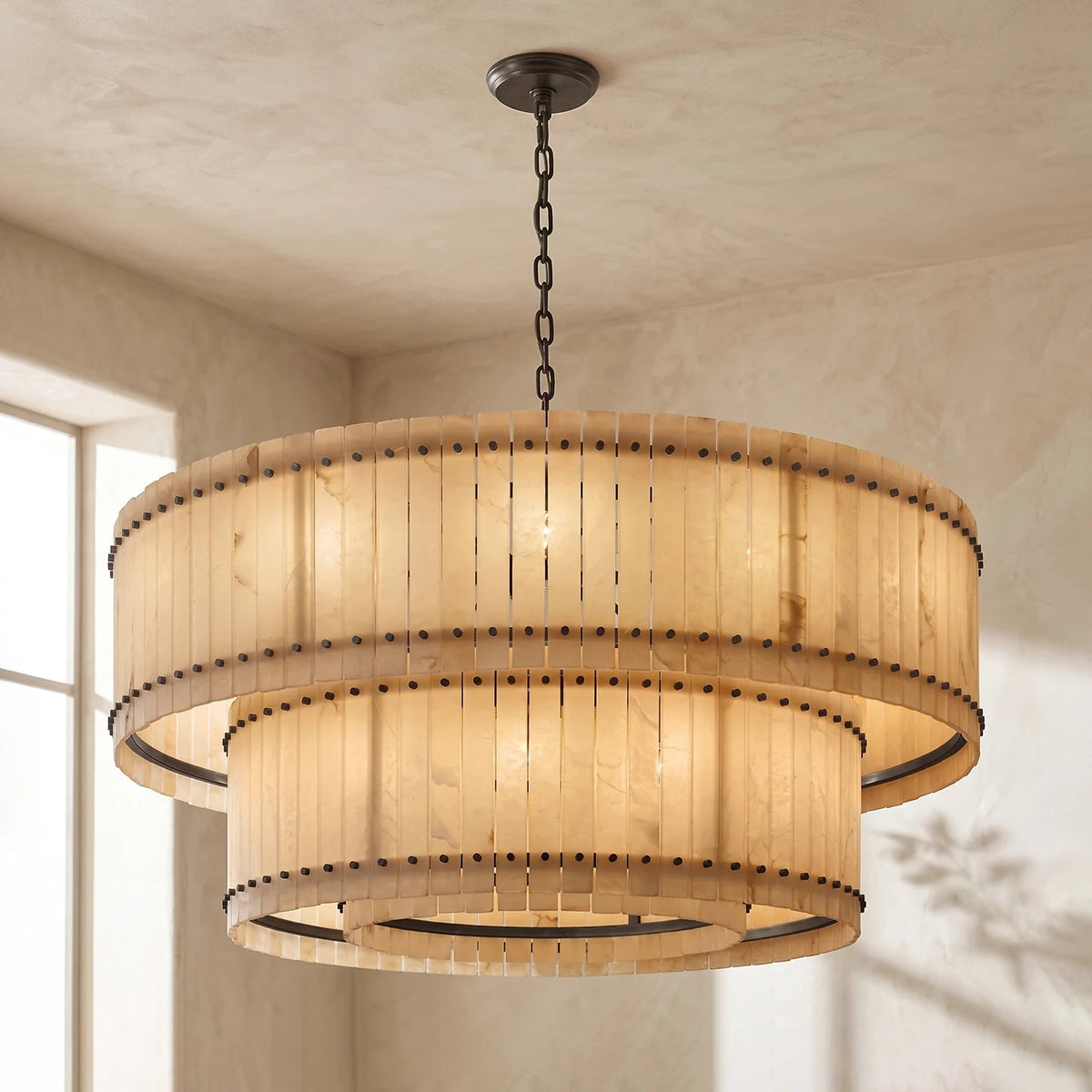 A two-tiered drum chandelier with a warm beige woven shade and dark metal hardware, hanging from a chain in a room with textured beige walls.