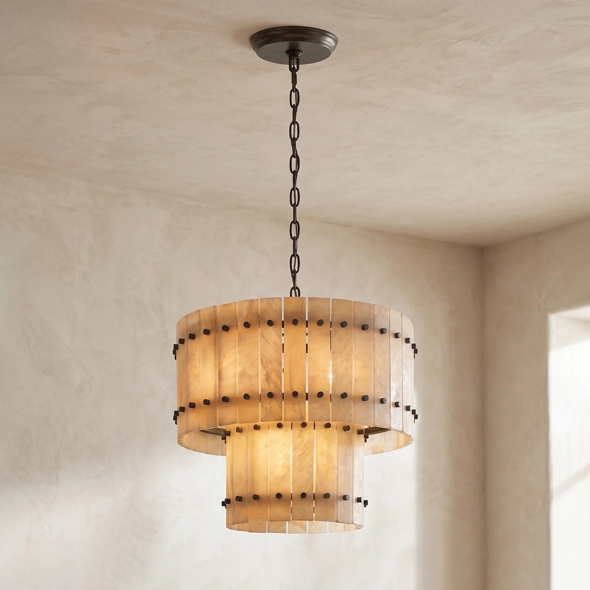 Illuminated two-tier chandelier with cream alabaster panels and dark metal fittings hangs from a dark chain in a neutral room.