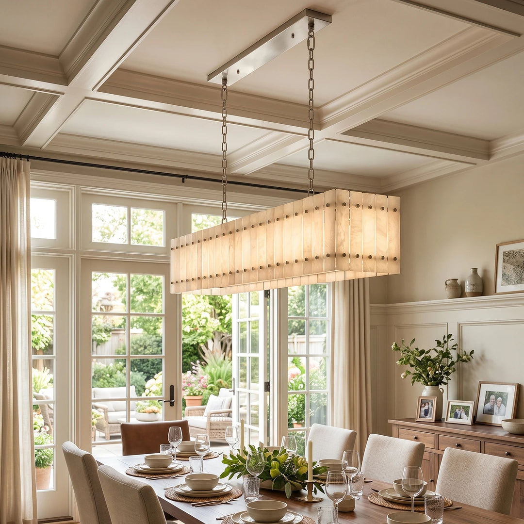 A rectangular chandelier with a warm beige pleated fabric shade and brass-toned hardware, hanging in a bright, elegant dining room with coffered ceilings and large windows.