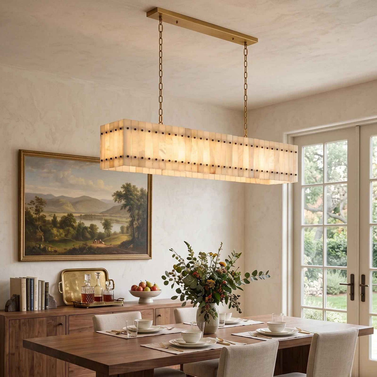 A linear alabaster chandelier with brass chains hangs above a wooden dining table, with landscape artwork behind it.