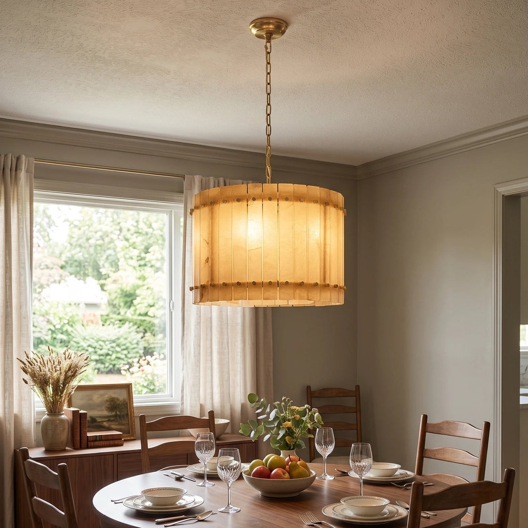 A cylindrical chandelier with glowing alabaster panels and a brass suspension chain hangs above a round wooden dining table.