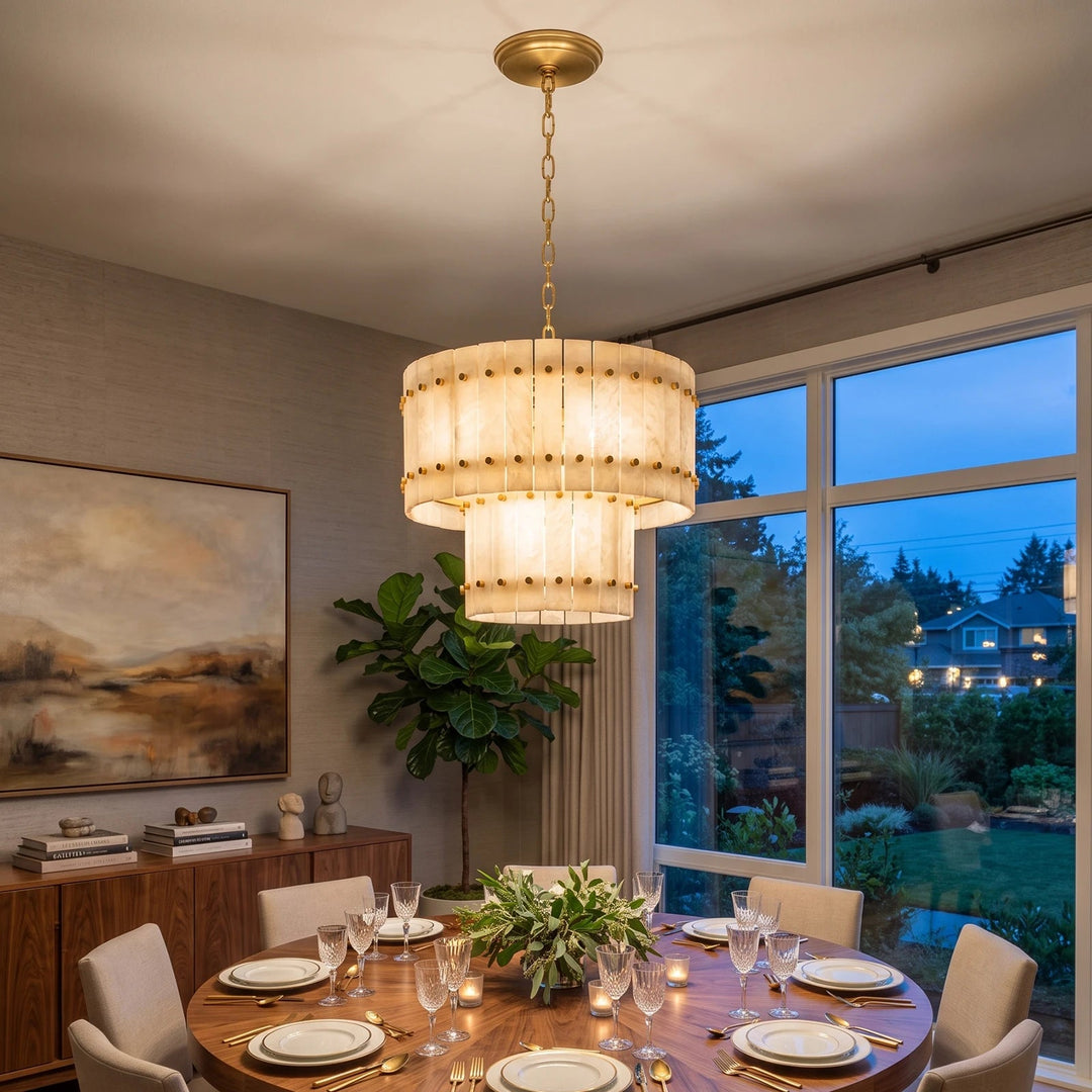 A modern multi-tier drum chandelier with a warm cream fabric shade and brass-toned metal hardware, hanging over a wooden dining table set for a dinner party in a contemporary dining room.