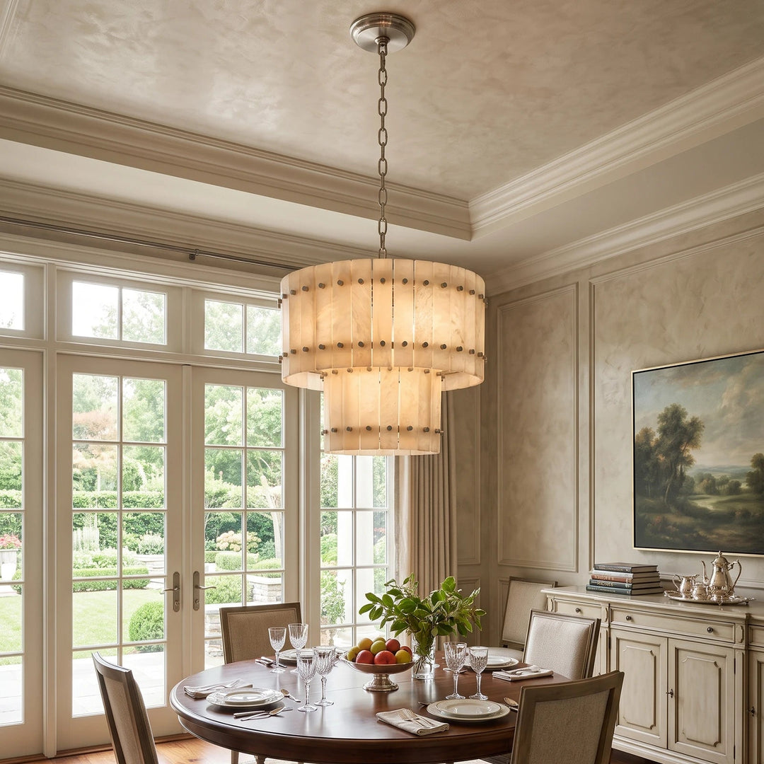 A large multi-tiered drum pendant chandelier with a warm beige woven shade and silver-toned metal hardware, hanging in a classic dining room with a wooden table set for dining and large windows showing a garden view.
