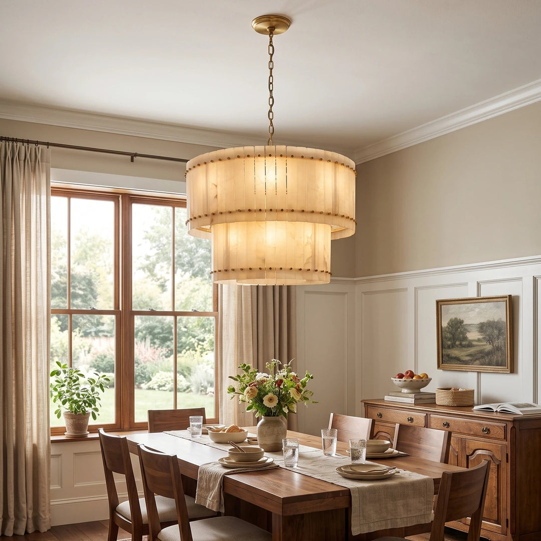 A large, tiered drum pendant chandelier with a warm beige fabric shade and brass-toned hardware hangs over a wooden dining table in a beautifully styled dining room.