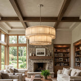 A large, multi-tiered drum chandelier with a warm beige woven or slatted shade hangs in a high-ceilinged living room with exposed wooden beams, a stone fireplace, and built-in bookshelves.