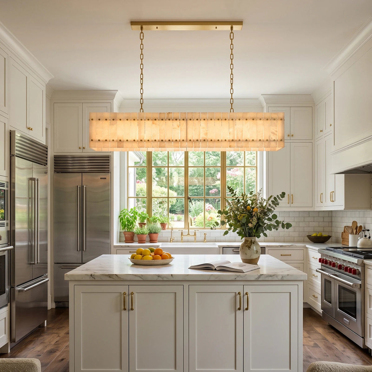 A linear chandelier with cream alabaster panels and brass chains hangs above a marble-topped island in a white kitchen.