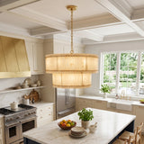A large cylindrical pendant chandelier with a warm beige woven shade and gold-toned hardware hangs over a marble kitchen island in a bright, modern kitchen with cream cabinets and stainless steel appliances.
