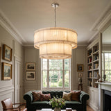 A large tiered drum pendant chandelier with a warm cream fabric shade and brass-toned metal accents, hanging in a classic, elegant living room with a green sofa and bookshelves.