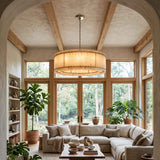A large cylindrical drum pendant chandelier with a warm beige woven or fabric shade hangs in a bright, sunlit living room with wood beams, a sectional sofa, and potted plants.